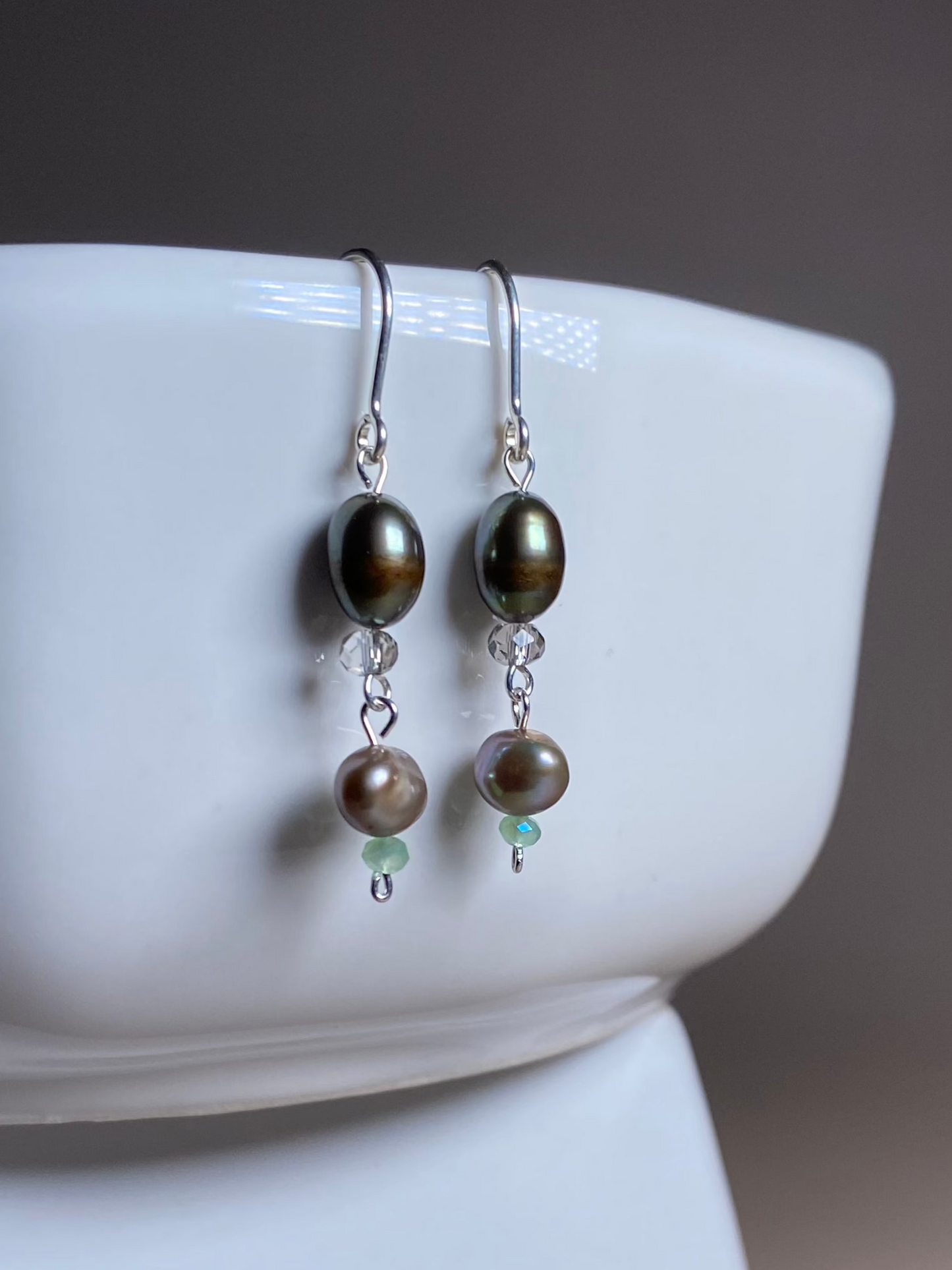 Erin Pearl Dangle Earrings gracefully hanging from the side of a white porcelain dish, showcasing their lustrous brown and grey pearls with shimmering green and silver beads and 925 sterling silver hooks.