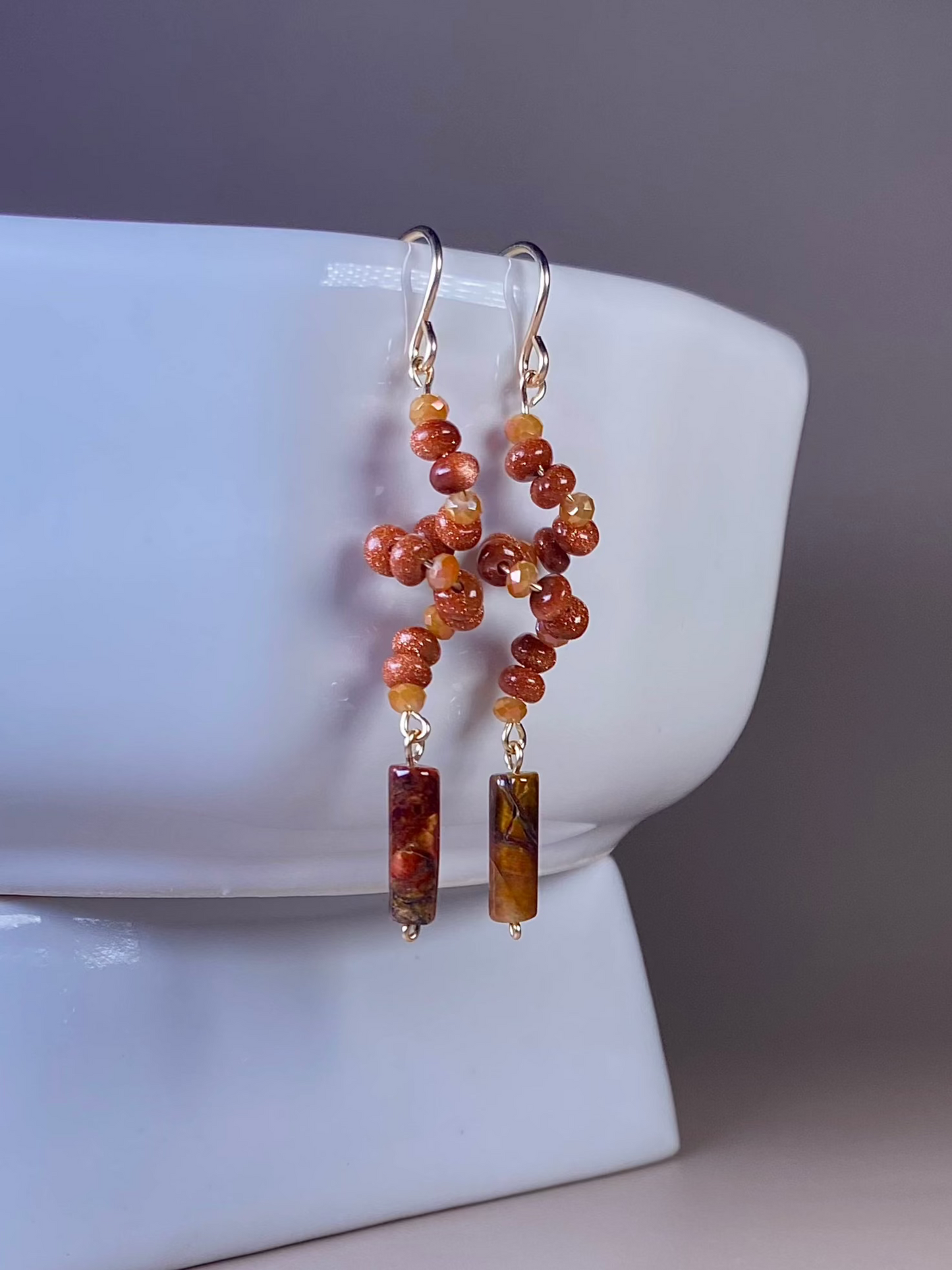 Francesca Moonstone Swirl Earrings, gracefully hanging from the side of a white porcelain dish, showcasing the golden moonstone swirl design with orange/gold glass beads and a pietersite bead on gold-filled hooks.