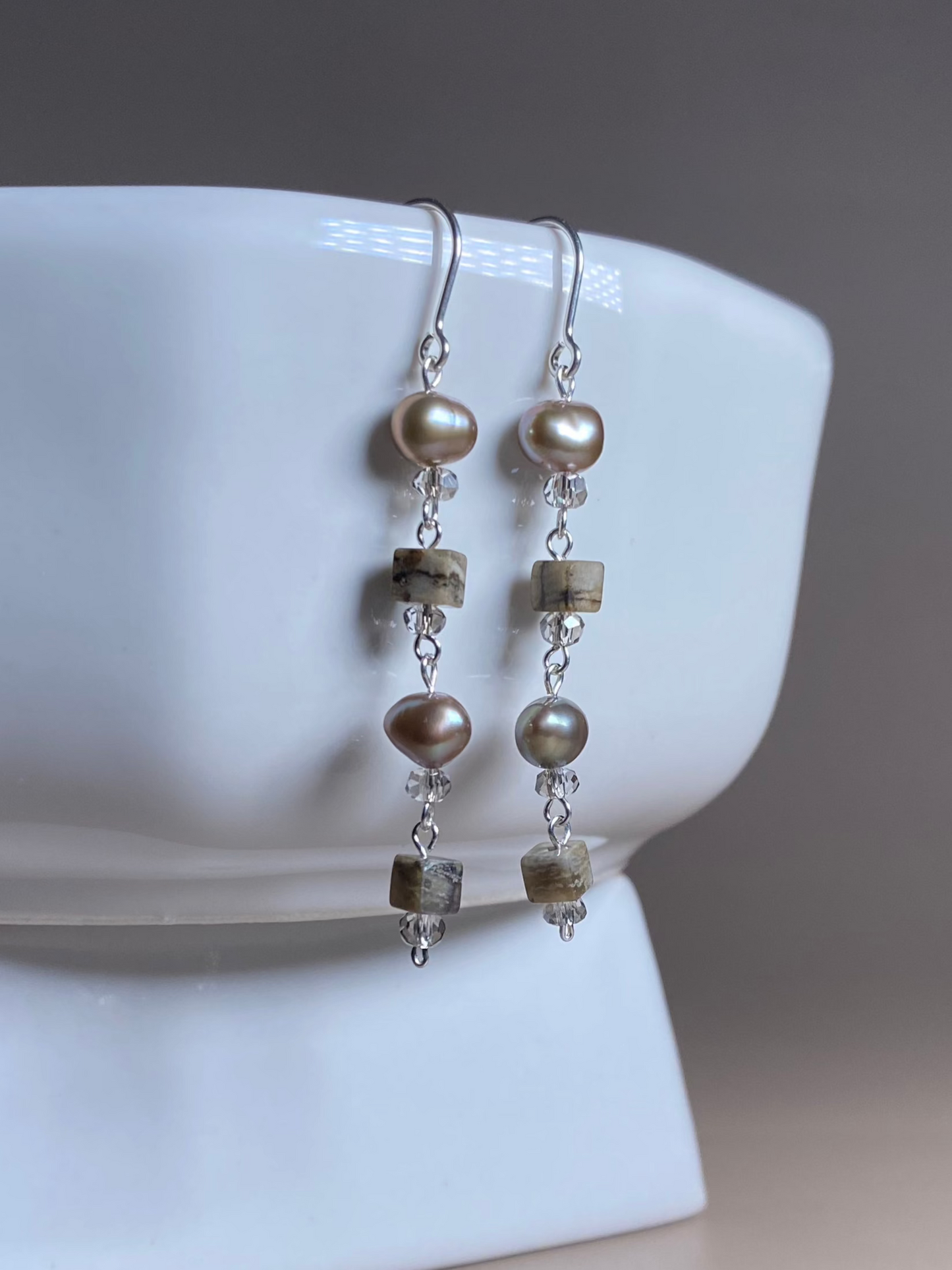 Veronica Pearl & Zebra Earrings with 6mm silver pearls and brown zebra cube beads, elegantly hanging from the side of a white porcelain dish, shining under the light.