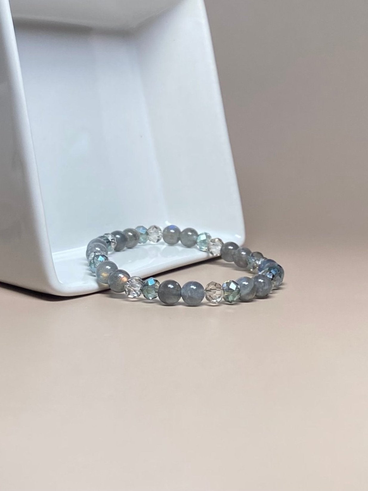 The Abby 6mm Labradorite Bracelet elegantly draped over a white porcelain container, showcasing its radiant A+ labradorite beads. The iridescent gemstones shine against the sleek, minimalist backdrop, enhancing the bracelet’s modern, luxurious look.