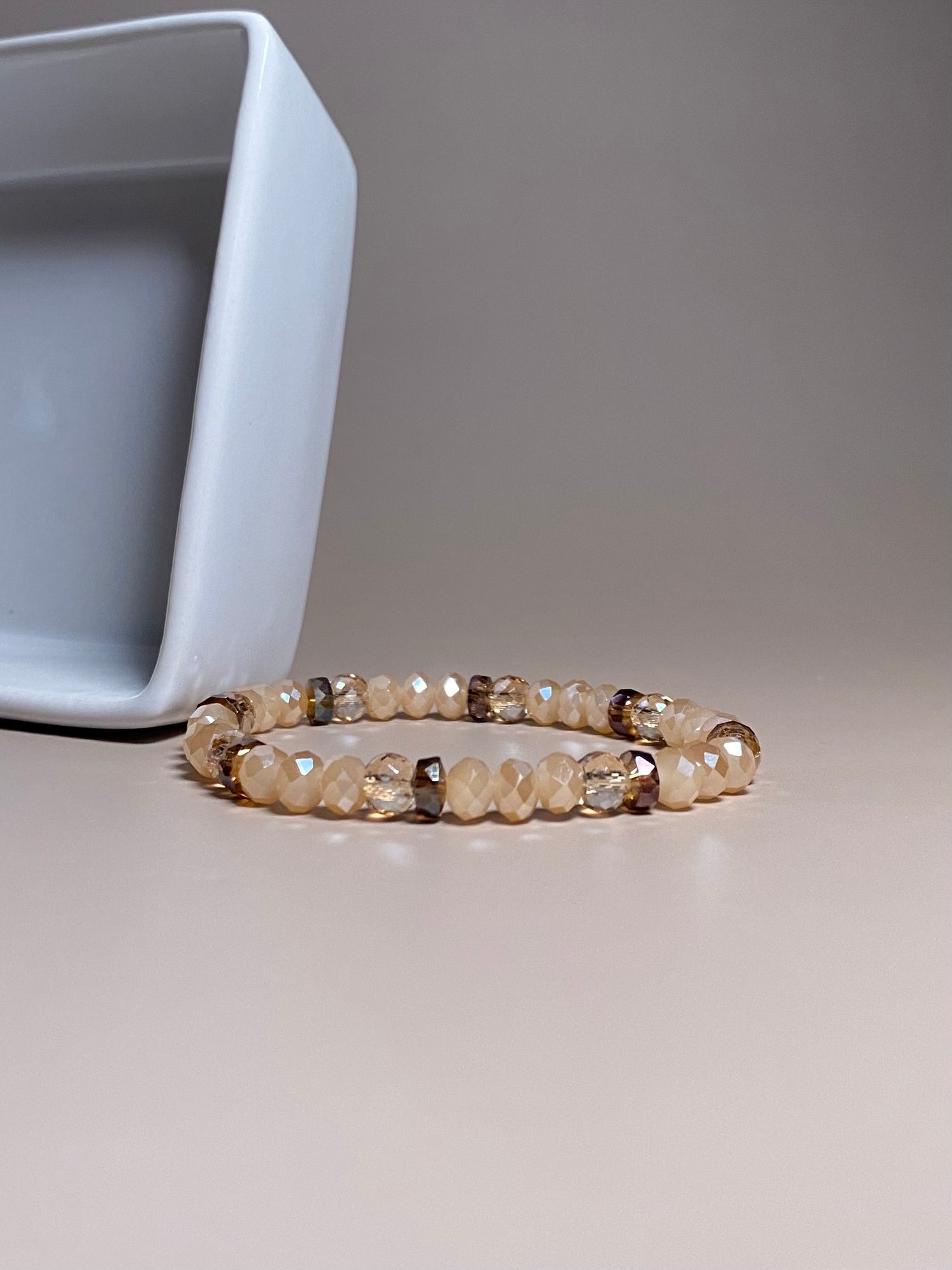 The Chelsea 6mm Cream Bracelet, adorned with 6mm shiny opaque cream beads and a mix of sparkly and brown beads, beautifully showcased in front of a square white porcelain container, highlighting its intricate design.