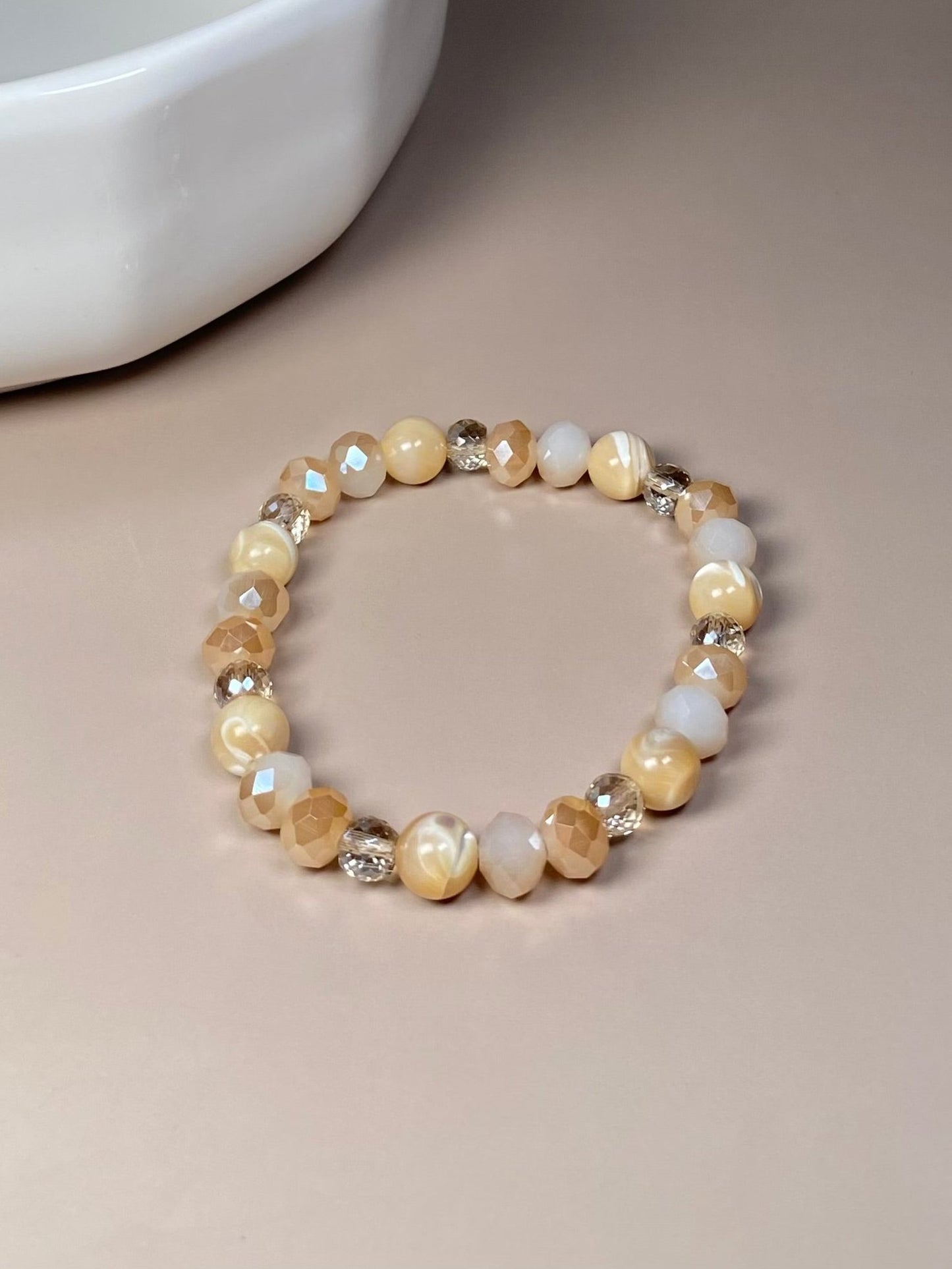 The Stella 8mm Mother of Pearl Bracelet elegantly positioned in front of a delicate porcelain dish, showcasing its combination of mother of pearl and shiny cream beads.